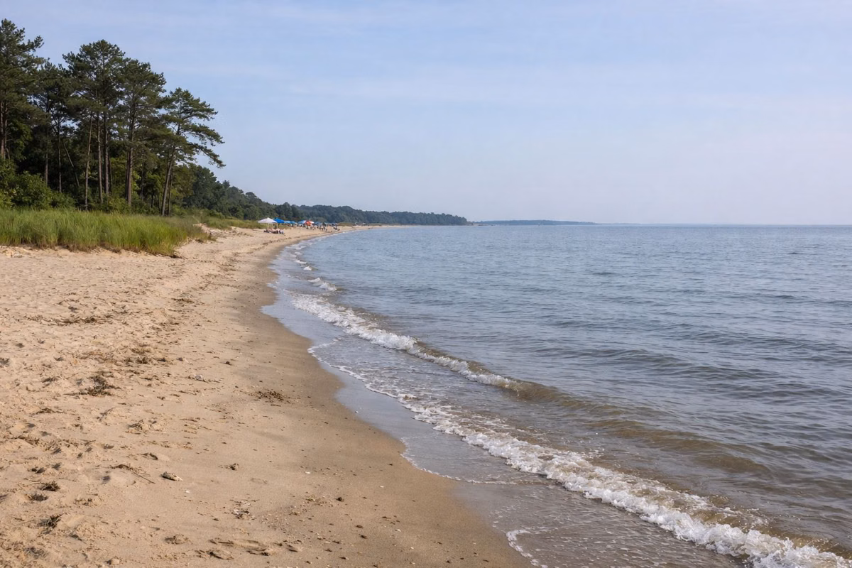 A wide sandy bay beach with gentle small waves, a few beach umbrellas far down the shore, open water to the horizon